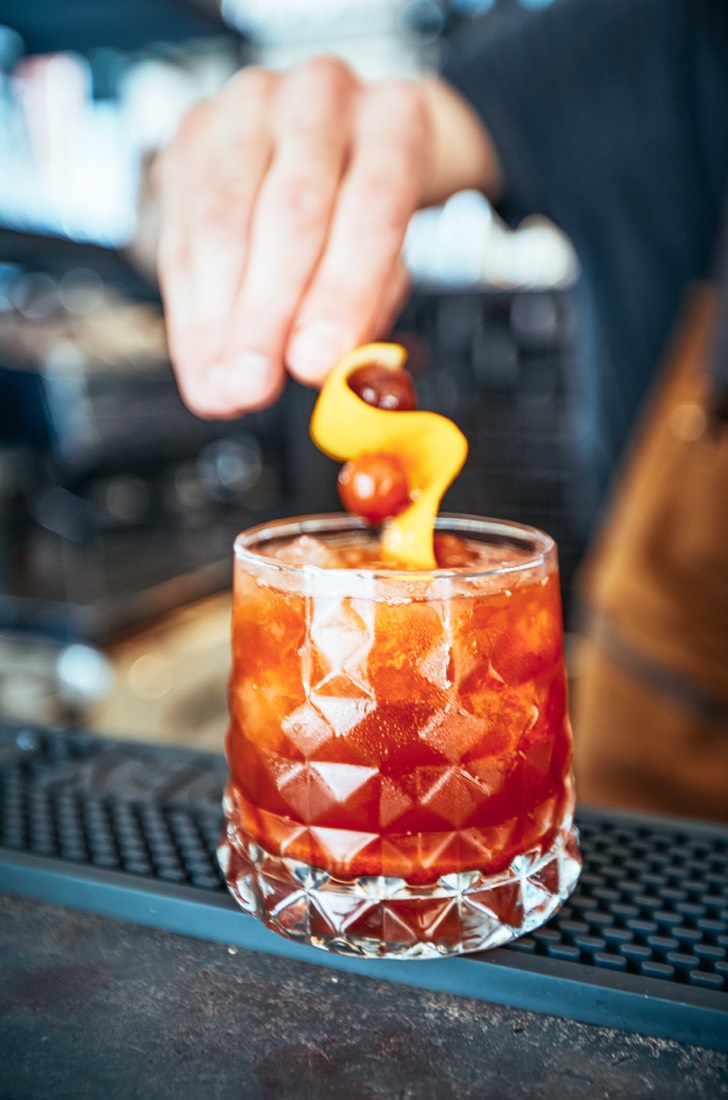 Un barman pose un zeste d’orange et deux cerises sur un cocktail servi dans un verre taillé
